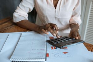 Person in a white blouse typing figures into a calculator, they know how to calcultate APS score and decide if they need a Matric rewrite, notebooks open on the desk as they work