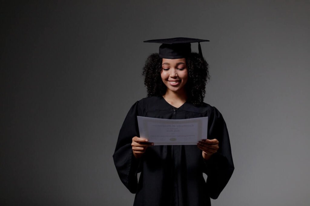 Student with curly hair, in graduation robe and hat, holding an original matric certificate, smiling from ear to ear